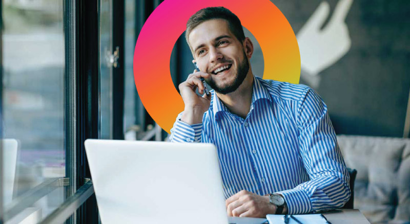A business professional talking into a mobile phone while sitting at a desk with an open laptop