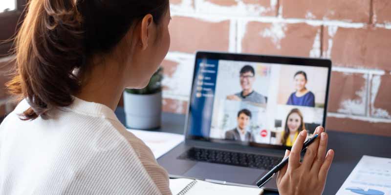 A person is attending a virtual meeting on their laptop while taking notes in a home office setting