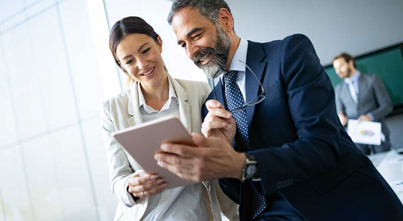 A man and a woman in business attire are reviewing a digital tablet together in an office environment