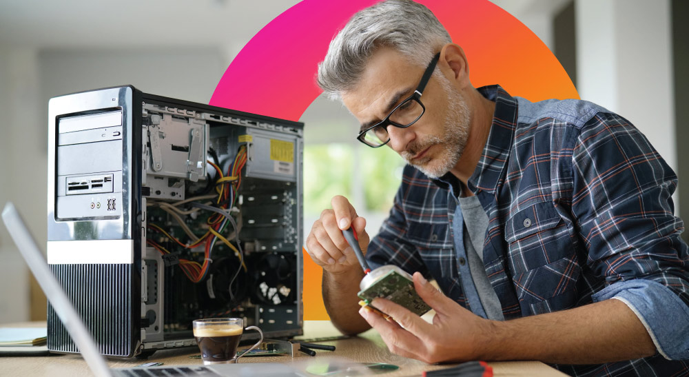 A person is repairing a computer hardware component with an open desktop tower and a laptop on the desk
