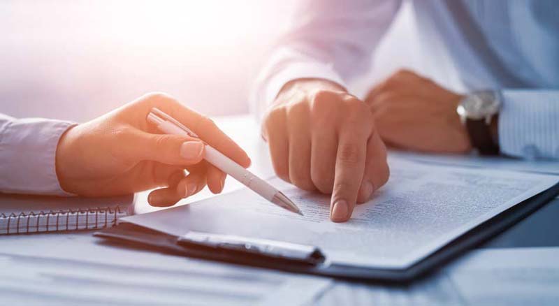 Two people's hands are shown in close-up, with one hand holding a pen and the other pointing at a document, suggesting a business setting