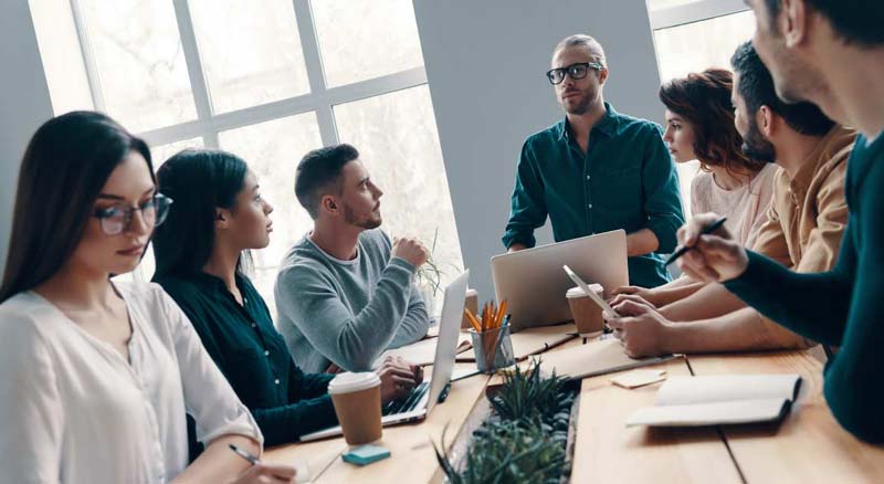 A group of seven individuals are engaged in a meeting, with one presenting while the others listen around a table