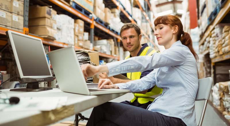 Two individuals working on laptops in a warehouse setting, surrounded by shelves stocked with various boxes.