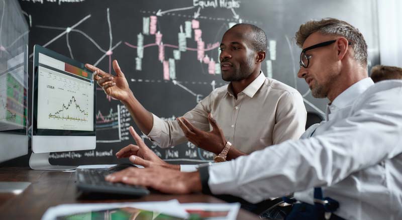 Two individuals in a business setting analyzing financial data on a computer screen, with one person pointing at the screen which displays a graph, and various documents on the desk in front of them.