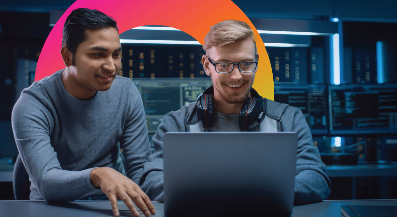 Two tech workers collaborating on a laptop in a blue-lit server room