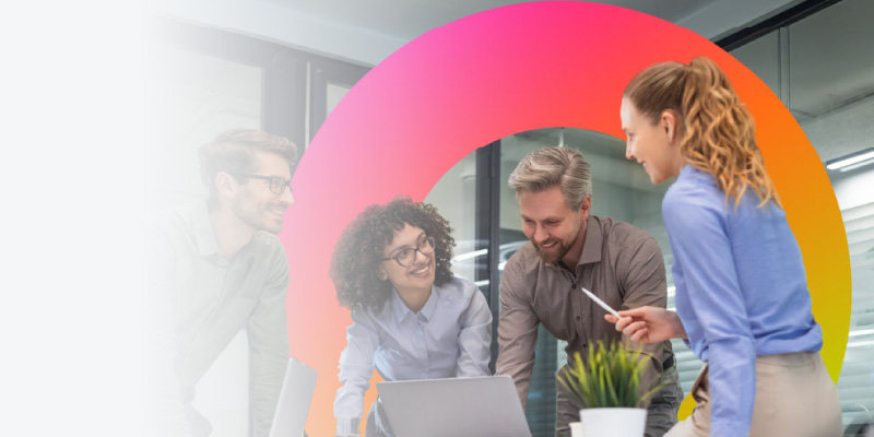A diverse group of four colleagues smiles and discusses around a laptop in a modern office.