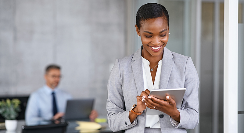 A person in business attire holding a digital tablet with another individual blurred in the background, indicating a professional setting