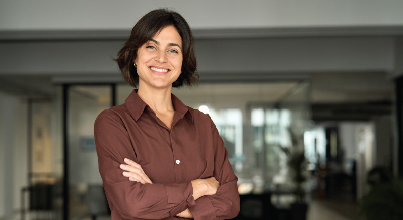 Woman with short brown hair in a brown blouse, smiling with arms crossed in a modern office.