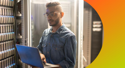 A man wearing glasses holds a laptop in front of a server, focused on his work in a tech environment