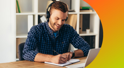 An student with headphones writing in notebook while using laptop for call