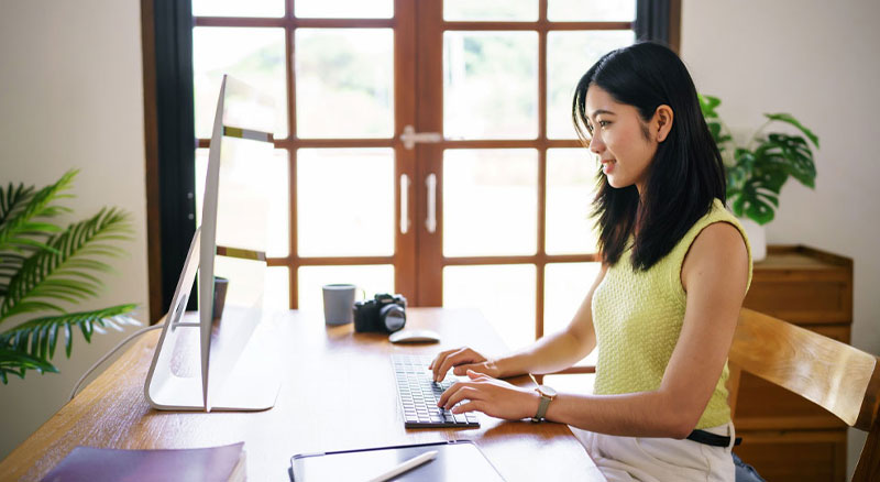 An individual sitting at a desk using a computer in a room with large windows and plants