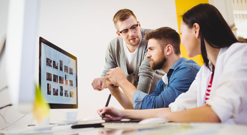 Three individuals collaborate around a computer monitor displaying various images
