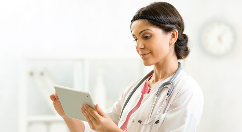 Healthcare professional in a white coat with a stethoscope around their neck, holding a tablet in a clinical setting.