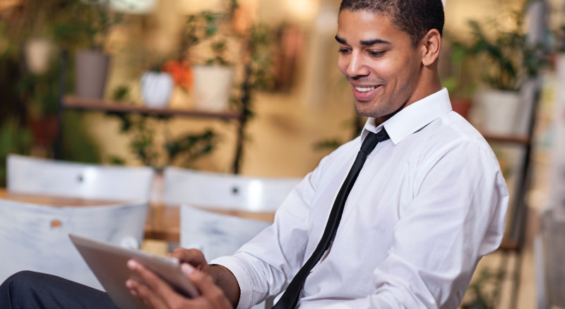 A person in formal attire using a tablet at a café, with a coffee cup on the table and empty chairs in the background