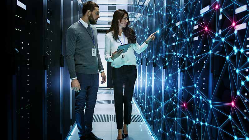 Two professionals examining a server room with illuminated network connections. The person on the right is holding a tablet and pointing towards the servers.