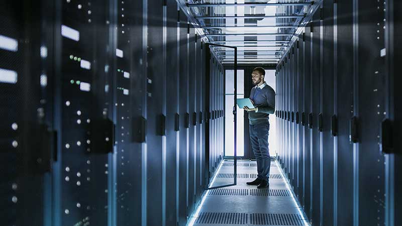 A person holding a laptop in a modern, illuminated data center with rows of servers.