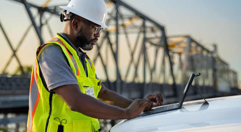 A government employee wearing a safety vest and hard hat uses a tablet outside