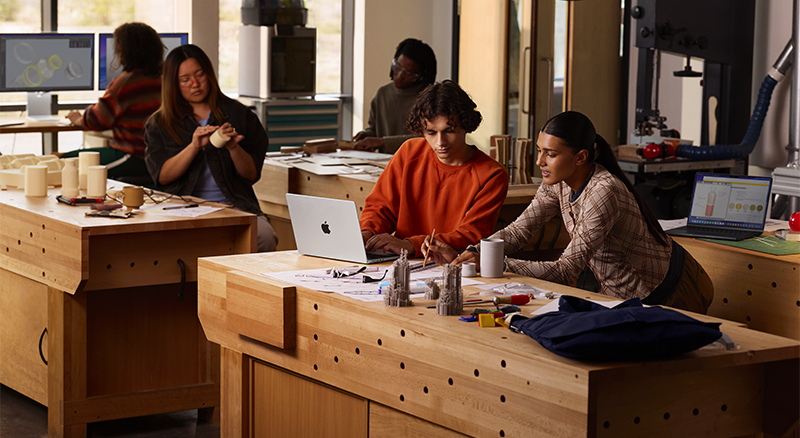 People working at wooden tables in a workshop with laptops, 3D models, and design tools scattered across the surfaces