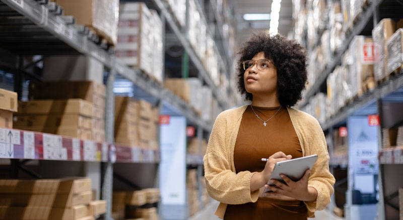 A person holding a tablet stands in an aisle between tall shelves in a warehouse