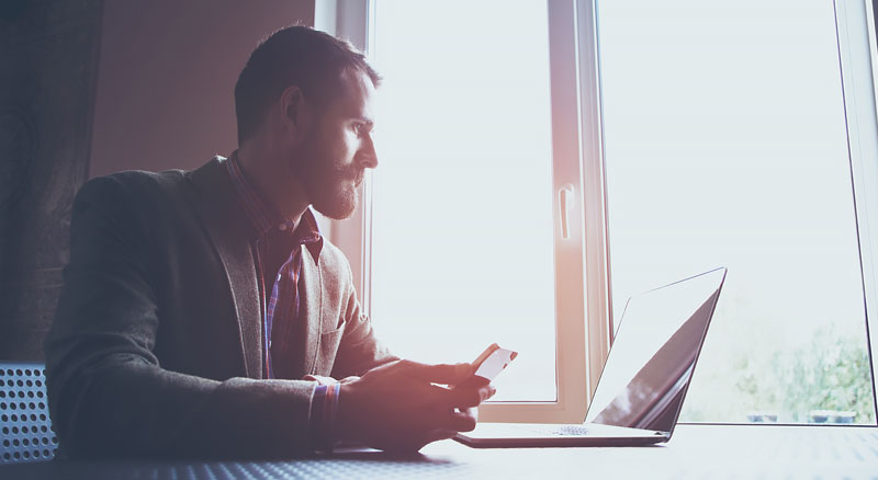 A person sitting by a window, using a smartphone with an open laptop beside them