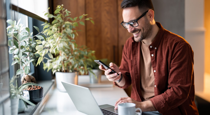 A person in a red shirt using a smartphone while working on a laptop, with a cup of coffee and potted plants nearby