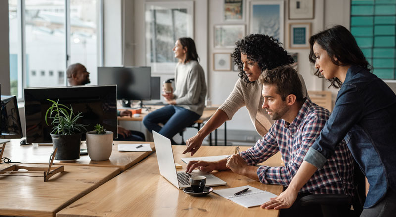 Three people are collaborating on a project over a laptop on a long wooden desk.