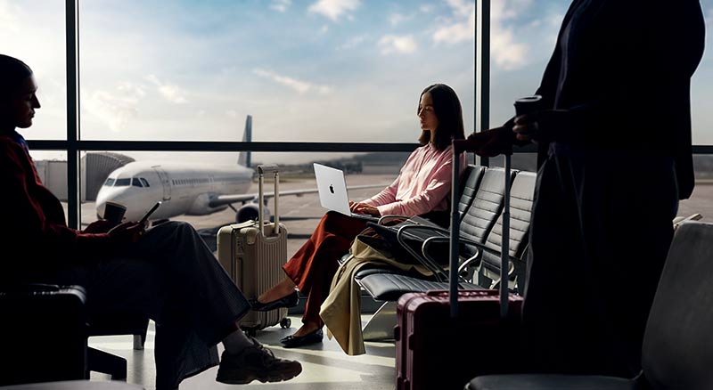 A person in a red shirt using a smartphone while working on a laptop, with a cup of coffee and potted plants nearby