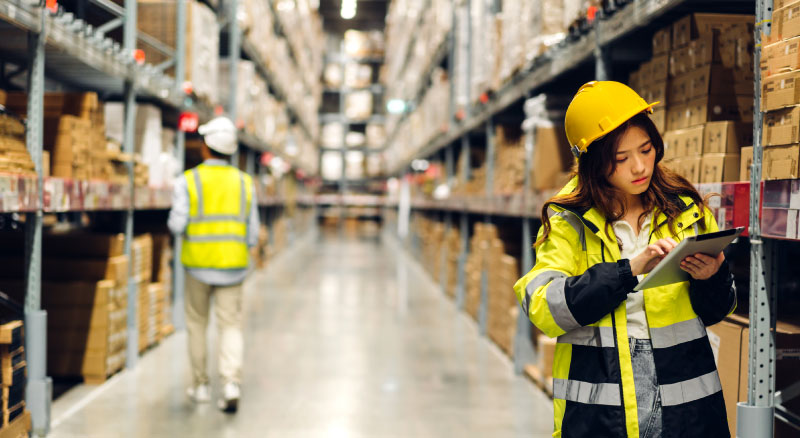 Two workers in a warehouse, one holding a clipboard and the other walking away