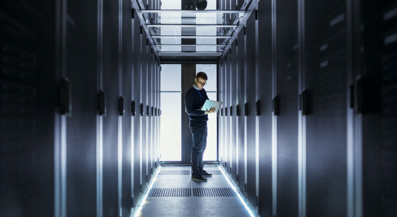 A person stands in a server room, holding a tablet while examining the equipment