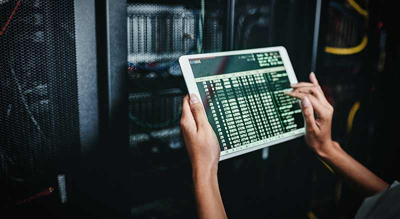 A person stands in a server room, holding a tablet while examining the equipment