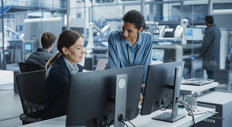Two people talking in an office setting while working on a computer