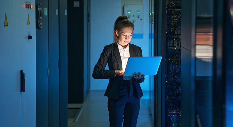 A person in a suit holding a laptop standing in front of a row of server racks