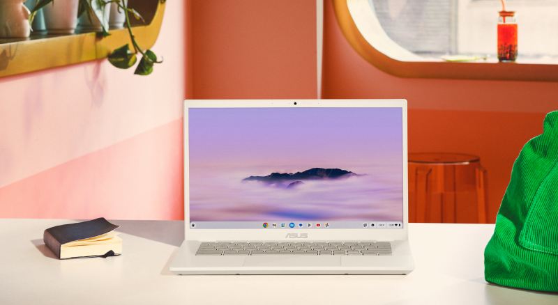 White ASUS laptop on a desk, displaying a screensaver of a cloud-covered mountain peak. The laptop is accompanied by a closed black notebook to its left and a green knitted bag to its right. The setting is against an orange wall, which has decorative items on a shelf.