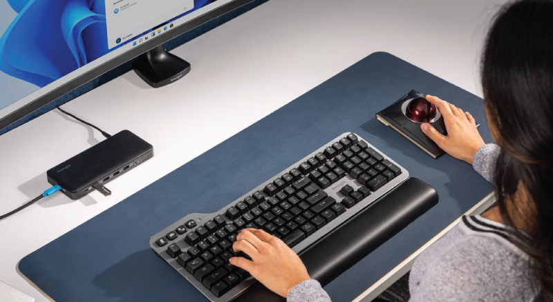 A woman uses a trackball mouse and keyboard on a desk with a large monitor. The setup has a sleek, organized look, conveying productivity.