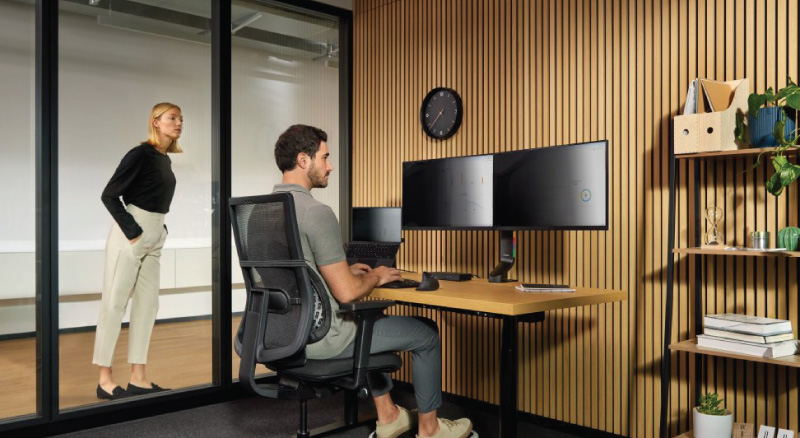 A man works at a desk with dual monitors in a modern office, viewed through a glass wall. A woman in business attire observes him. A clock and shelf with plants are visible.