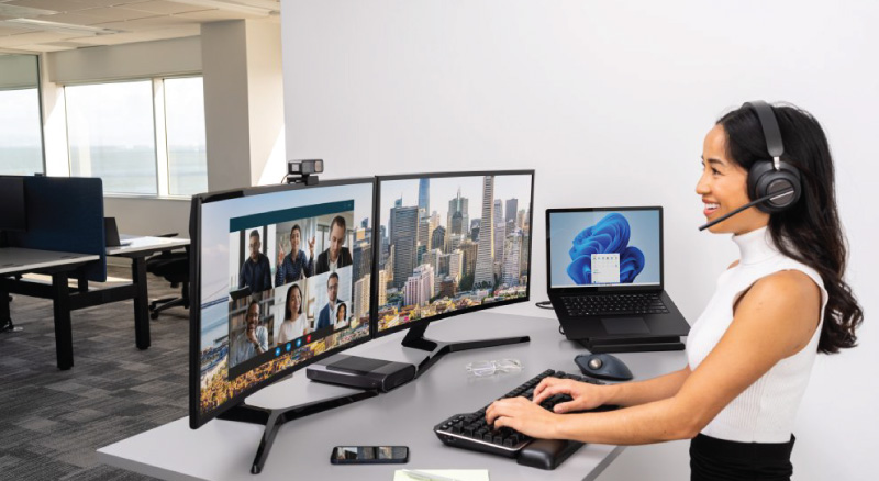 A woman in an office wears a headset, smiling while working on dual monitors displaying a video call.