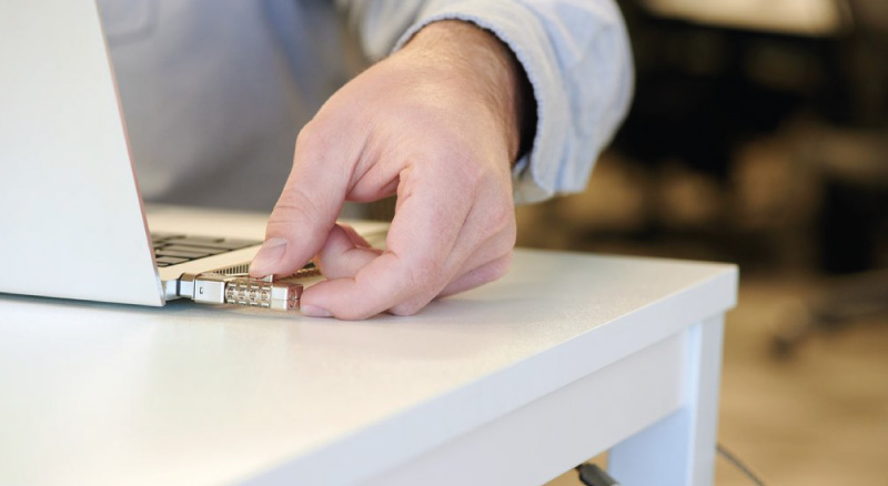 A person inserts a gold USB stick into a laptop on a white desk in an office. A cable secures the desk leg. The scene conveys a sense of caution and security.