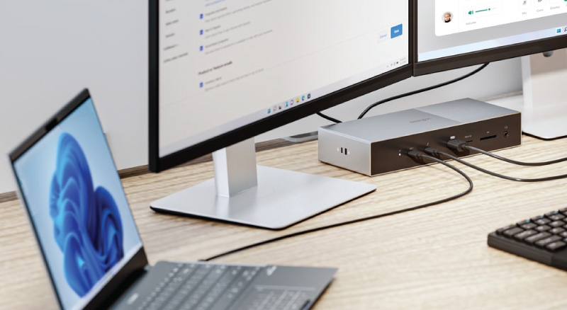 A modern office desk features a laptop displaying a blue abstract design, two large monitors showing text and video calls, and a docking station connecting them.