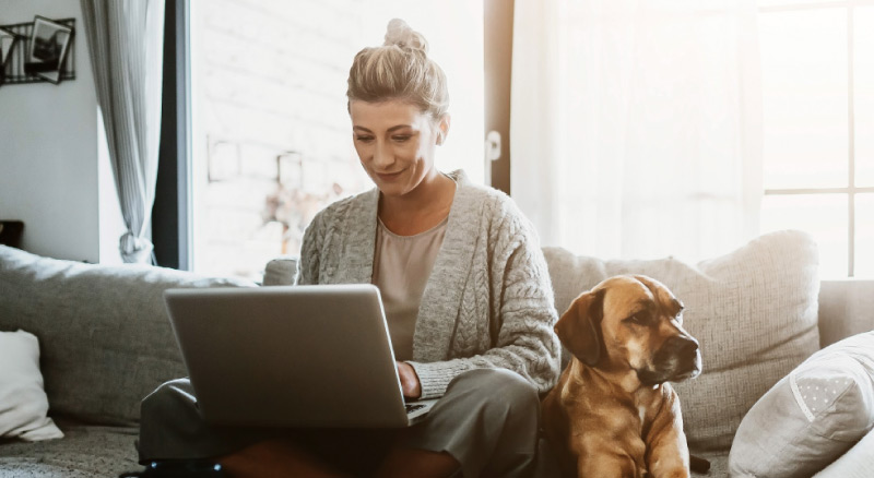 Woman at home sitting on a couch with her dog while on a laptop