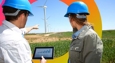 Two engineers in hardhats look at windmills while reviewing data displayed on a digital tablet