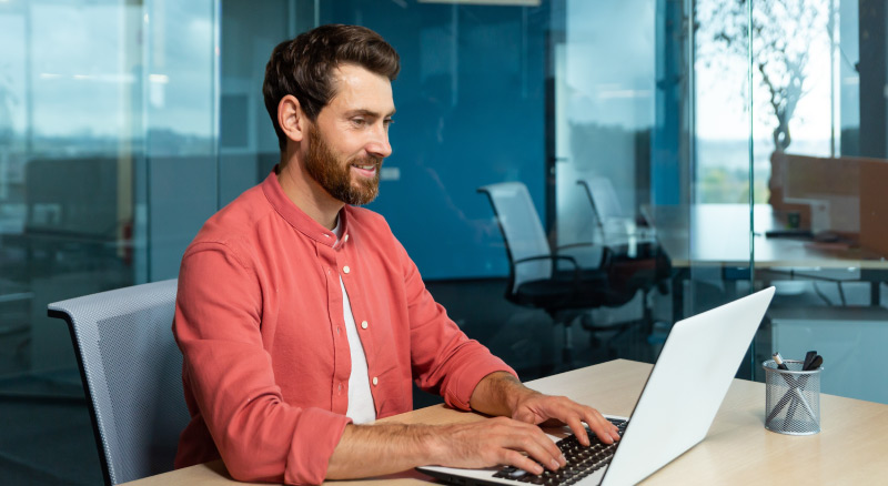 Young male student in a red shirt. Sitting at the desk in the office, studying online on a laptop.