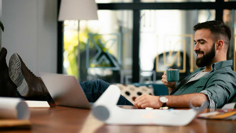 Man rests his feet on a desk beides his laptop while drinking coffee