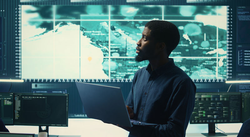 Tech employee using laptop in office surrounded by monitors