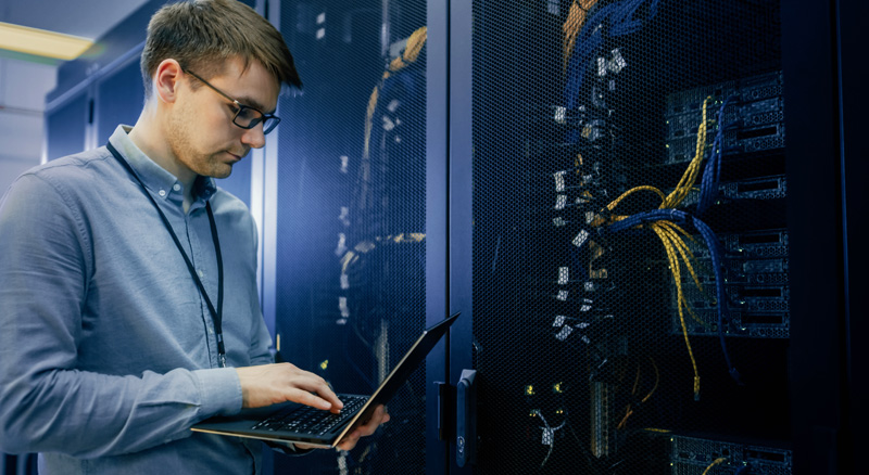 A person is using a laptop while standing next to open server racks with visible cables