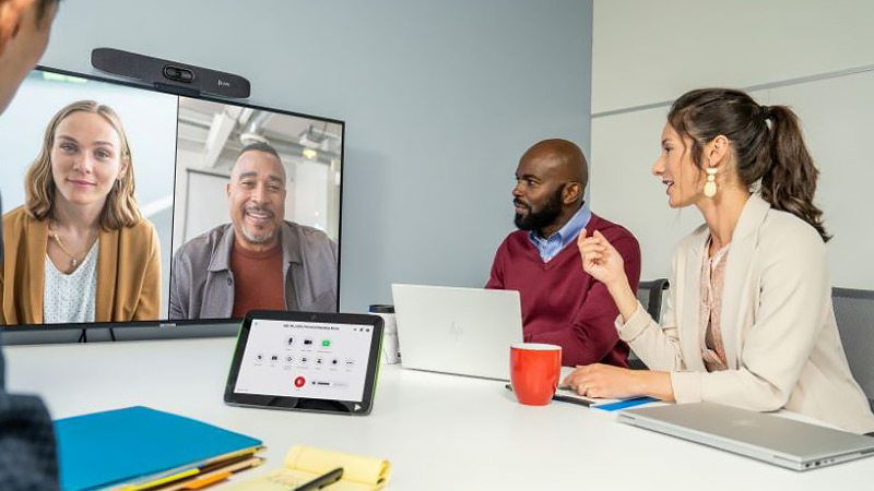 Employees gather at a table as they talk to two people remotely displayed on a television