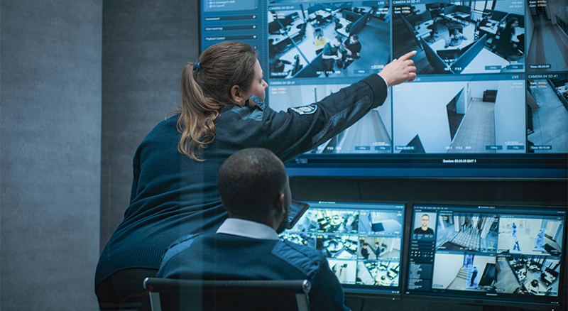 A woman gestures towards a security monitor with several screens, highlighting various data points or visuals.