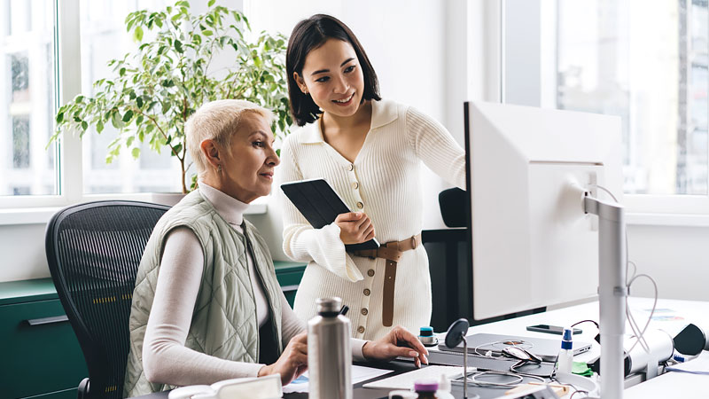 A woman assists her coworker while pointing at desktop monitor