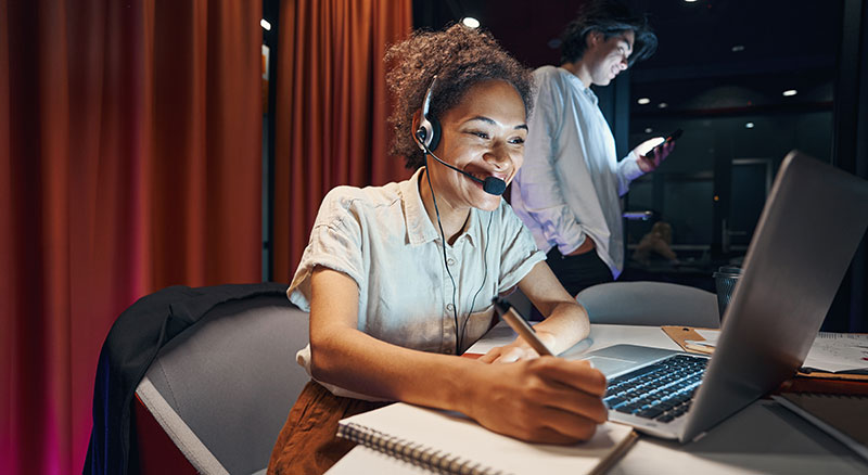 Person on a headset using a laptop and writing on a notepad