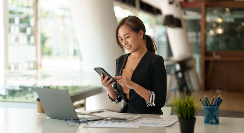 A person in a business suit sitting at a desk with a laptop, documents, and office supplies, holding and looking at a smartphone.