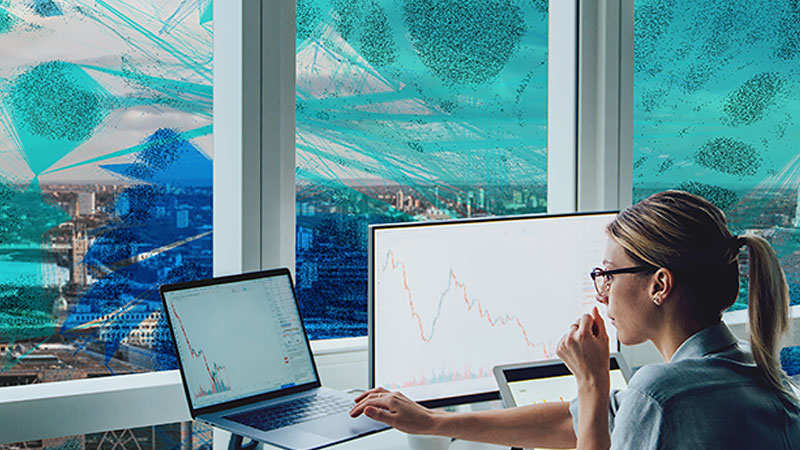 A woman works between a tablet, laptop, and computer monitor infront of a city skyline